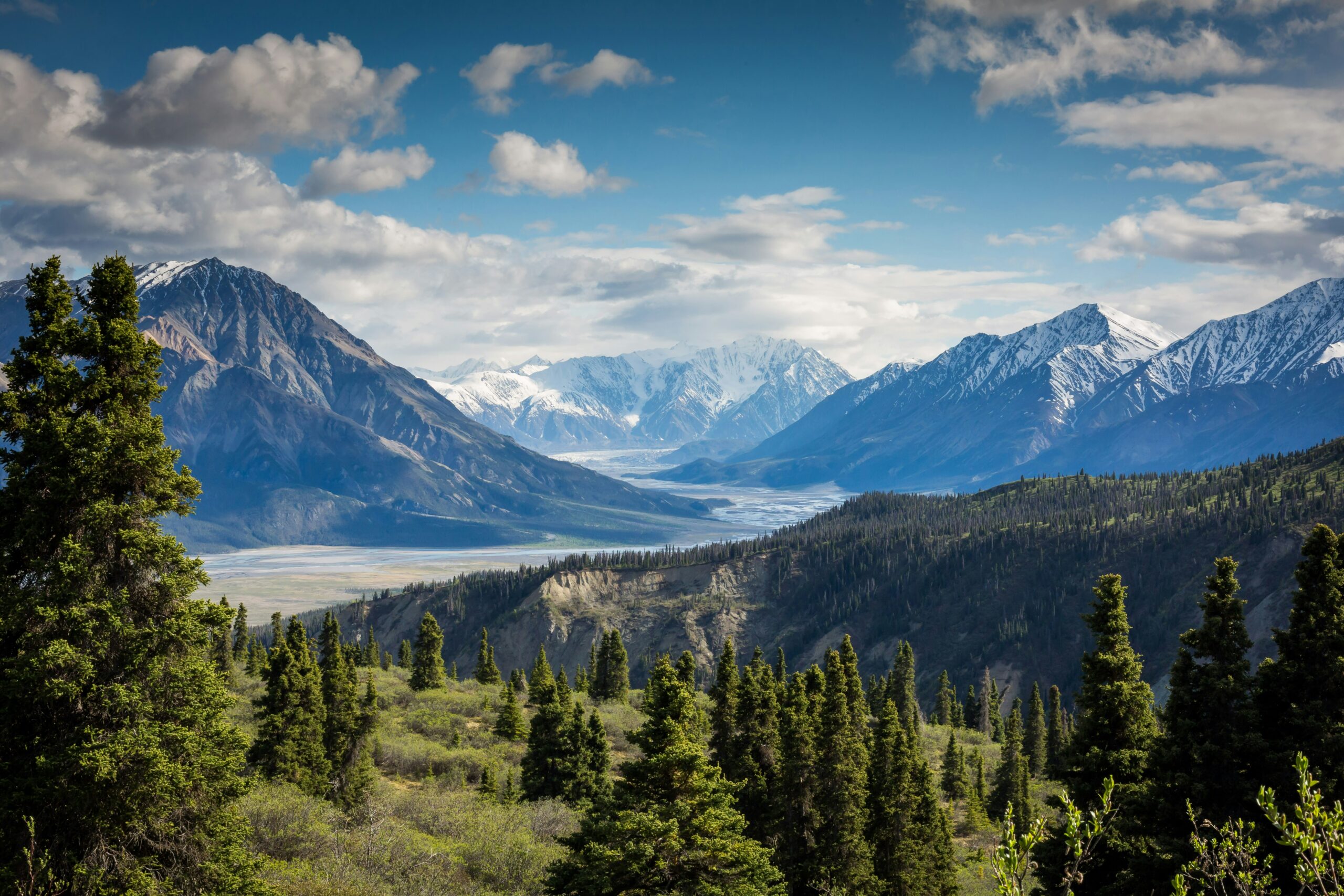 Mountain landscape with trees, snow-capped peaks, and cloudy sky.