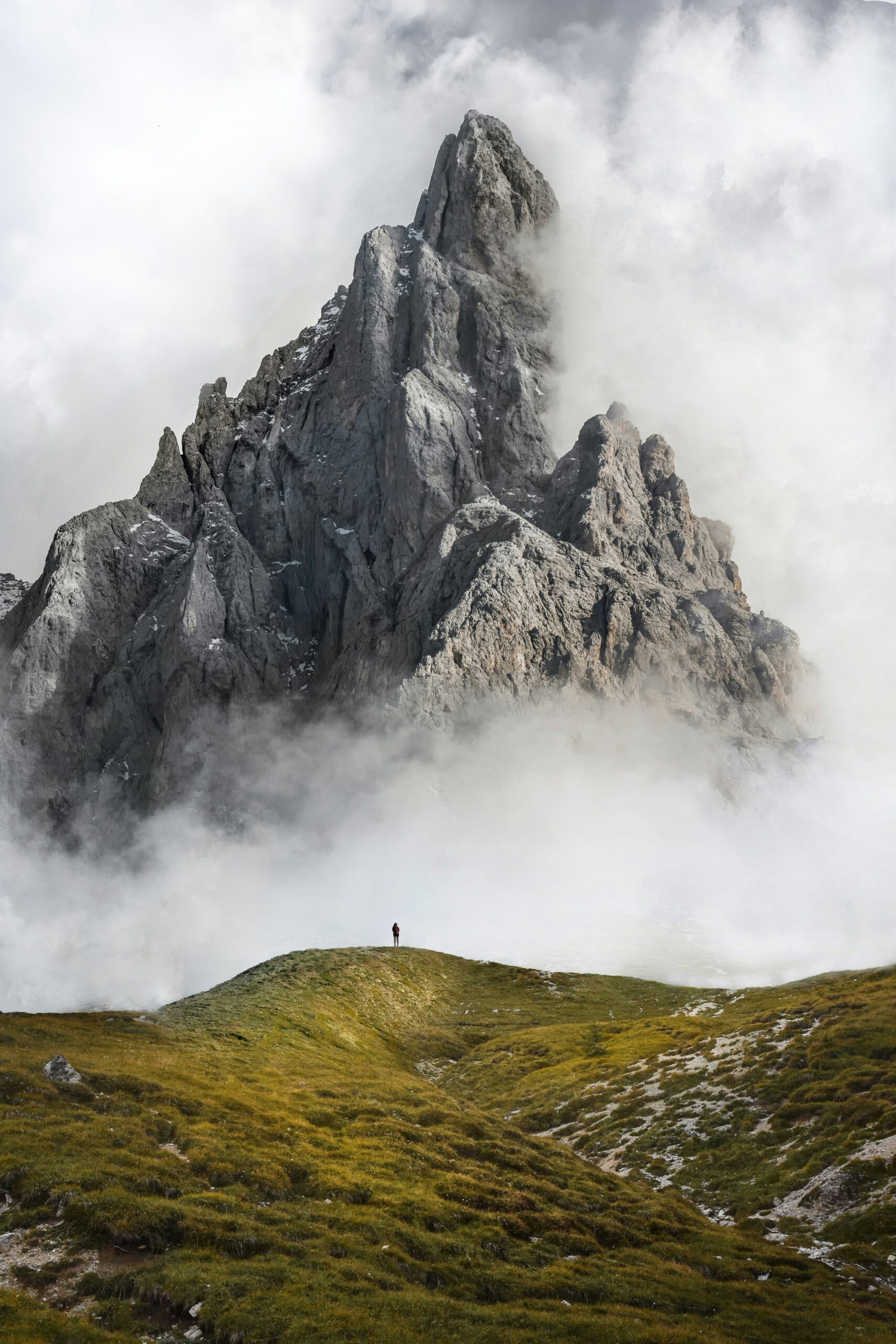 A person stands on a grassy hill below a misty, jagged mountain peak.