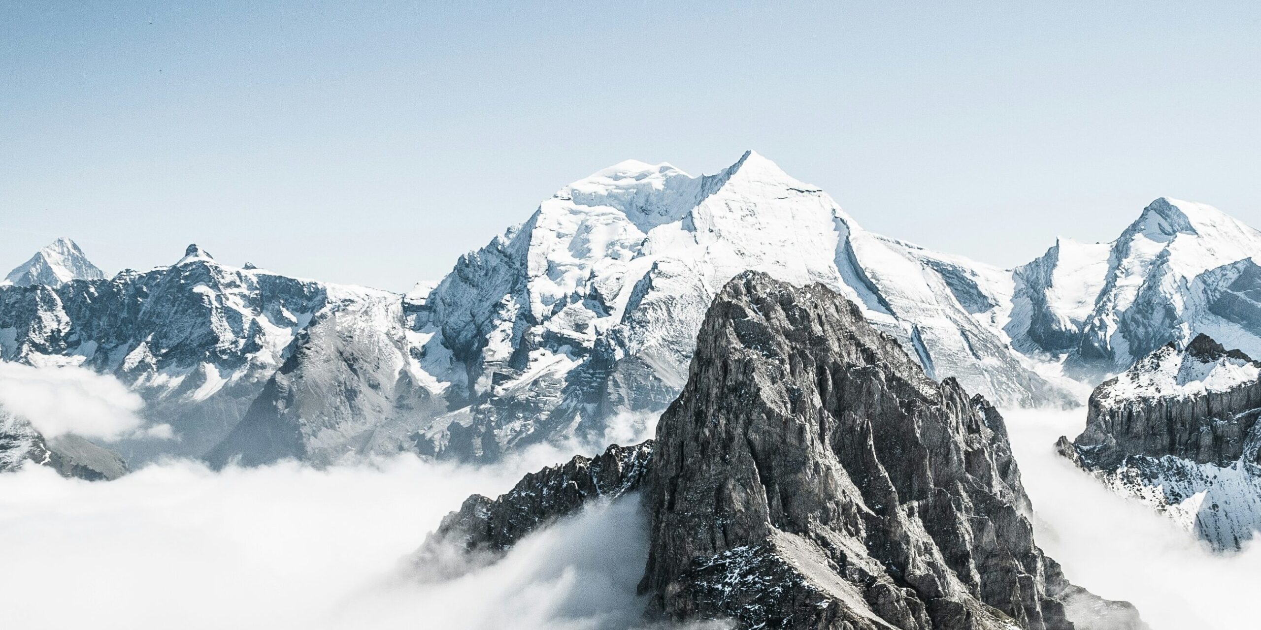 Snow-covered mountain peaks under a clear blue sky, surrounded by clouds