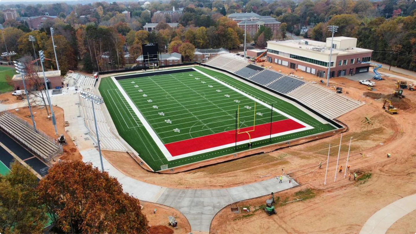 Aerial view of a football stadium under construction with a green field, red end zones, and a large adjacent field house