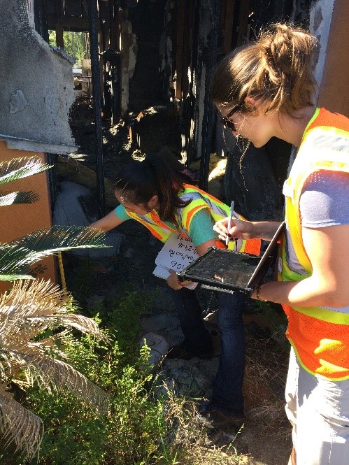 Laura Szczyrba of Dewberry’s Fairfax, Va., office enters damage assessment information on a tablet while Anne Lee, based in Gainesville, Va., searches for a water line to indicate the height of the flooding along a severely damaged home’s exterior.